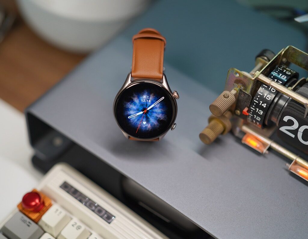 A round smartwatch with a brown leather strap sits on a gray surface next to a vintage clock radio. The smartwatch's face displays a digital clock and a colorful background