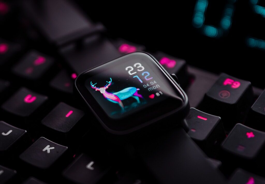 A black smartwatch with a dark background and a neon deer design sits on a keyboard. The smartwatch displays the time, date, and battery level.