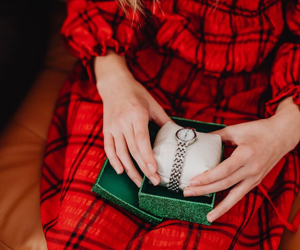 A person wearing a red plaid dress opens a green gift box to reveal a silver watch